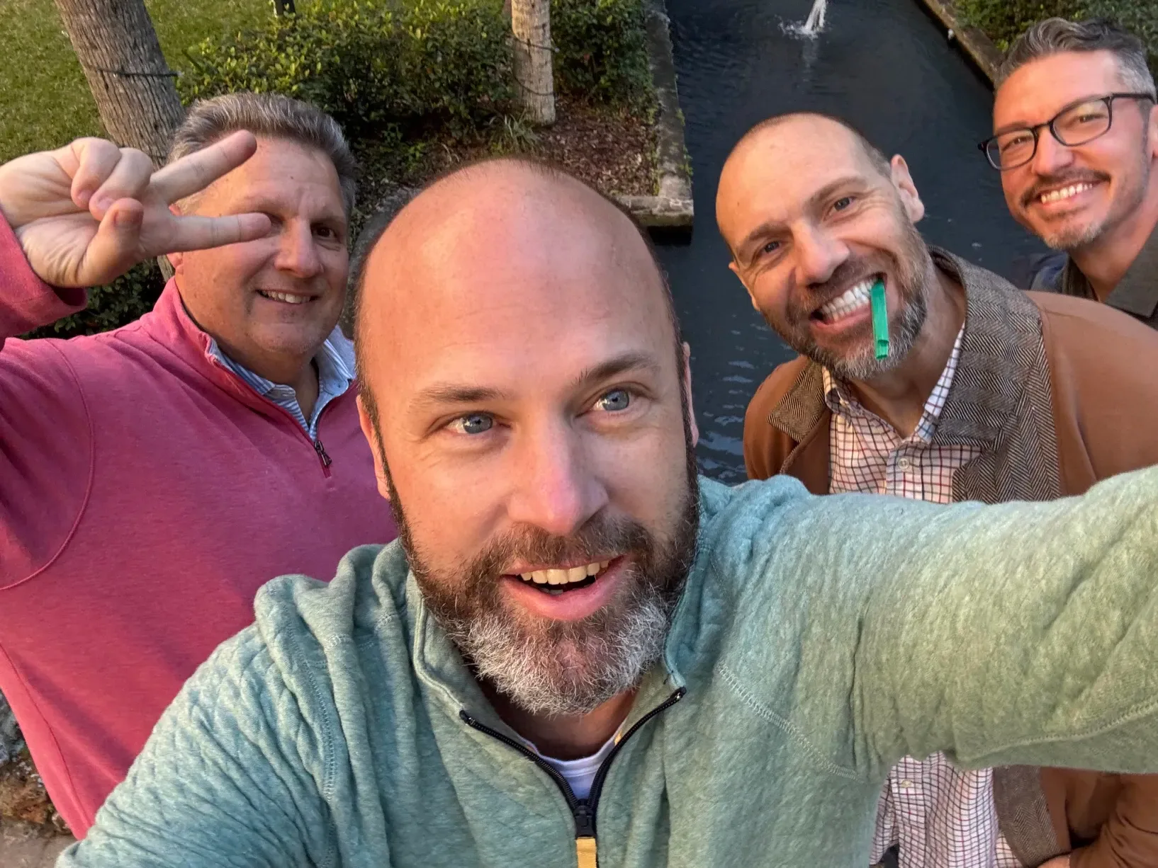 Pat taking a selfie with three smiling friends beside a canal; one holds up a peace sign and one has a green straw in his mouth