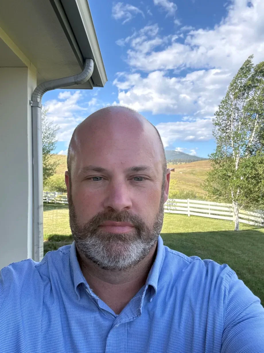 Pat, a bald man with a full beard in a blue checked shirt, in front of a white fence, green field, and Montana hills